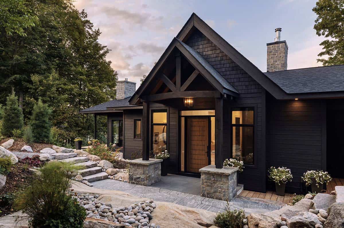 Dark-clad cottage front entrance with timber frame porch, stone pillars, and natural landscaping