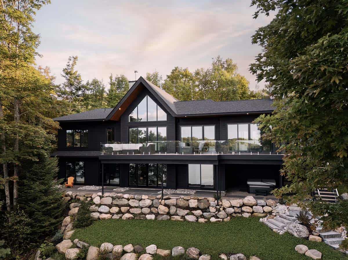 Two-story black cottage with large gabled windows, glass balcony railing, and stone retaining wall