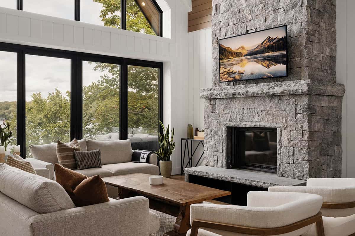Living room with gray stone fireplace tower, wall of black-framed windows, and neutral upholstered seating