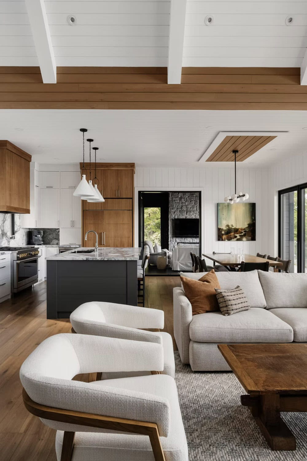 Bright open-plan living space with wood ceiling beams, boucle chairs, and kitchen island beyond