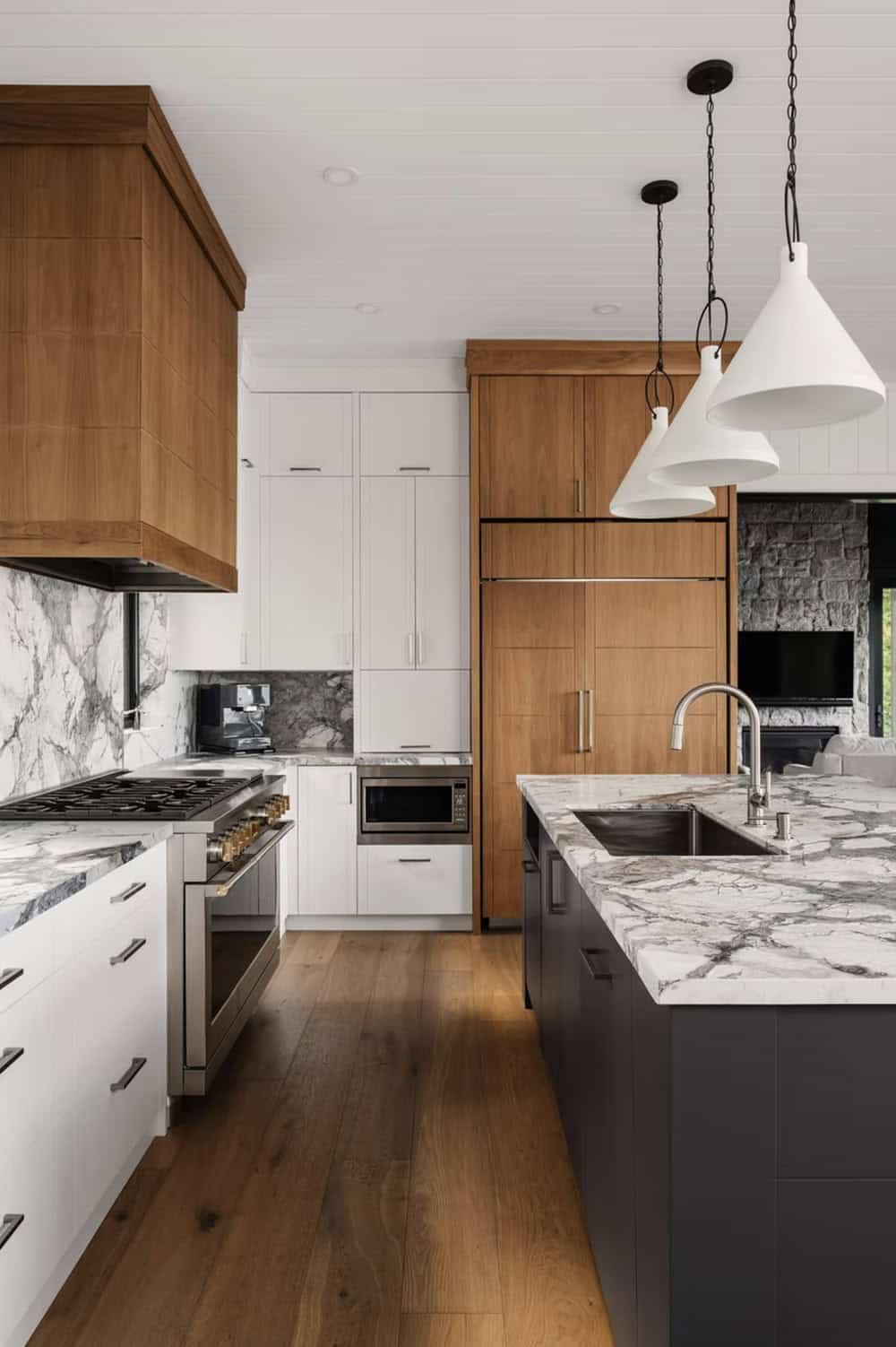 Kitchen galley view with gas range, marble surfaces, wood hood, and white pendants over dark island