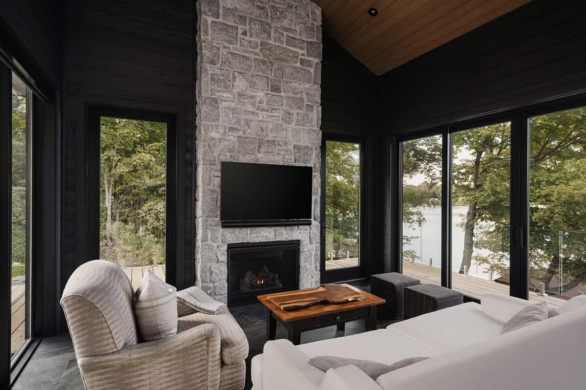 Dark sunroom with stone fireplace, wall of windows overlooking lake, wooden ceiling, and cozy seating
