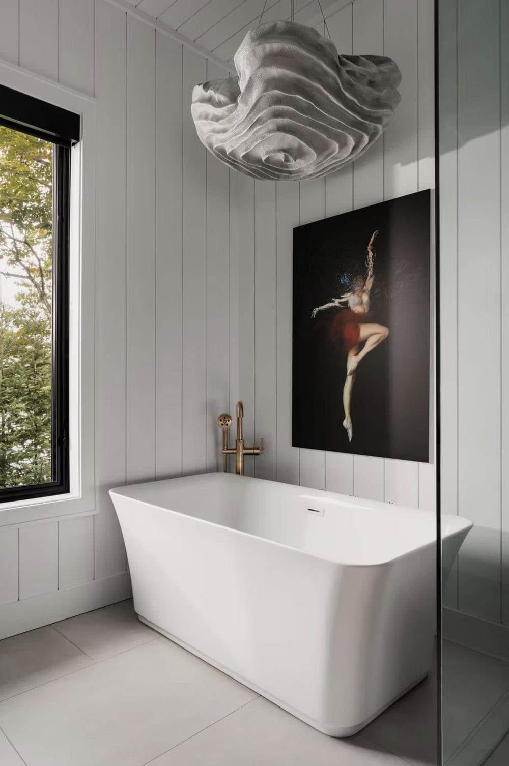 Freestanding white soaker tub with brass floor faucet, sculptural pendant light, and nature artwork above