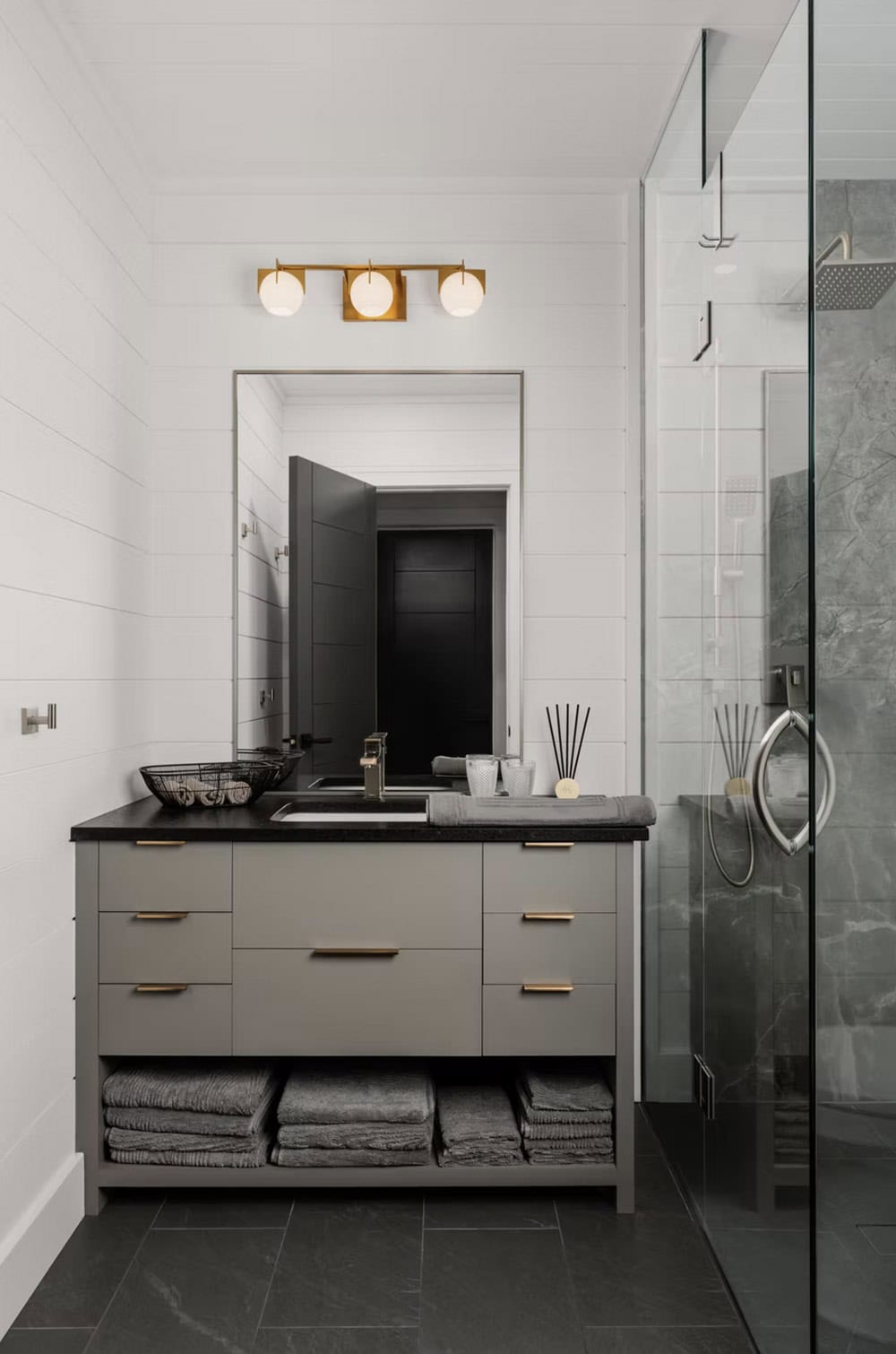 Guest bathroom vanity with taupe drawers, black countertop, brass globe lights, and glass shower