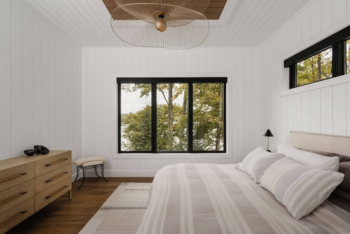 Bright guest bedroom with striped bedding, wood dresser, wire pendant light, and black-framed forest windows