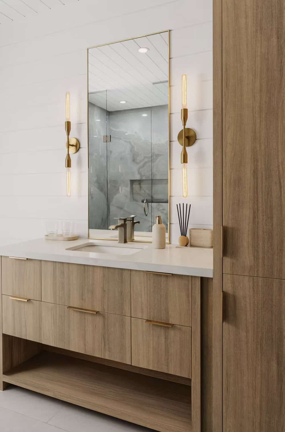 Bathroom vanity with light wood cabinetry, brass sconces, gold-framed mirror, and marble shower beyond