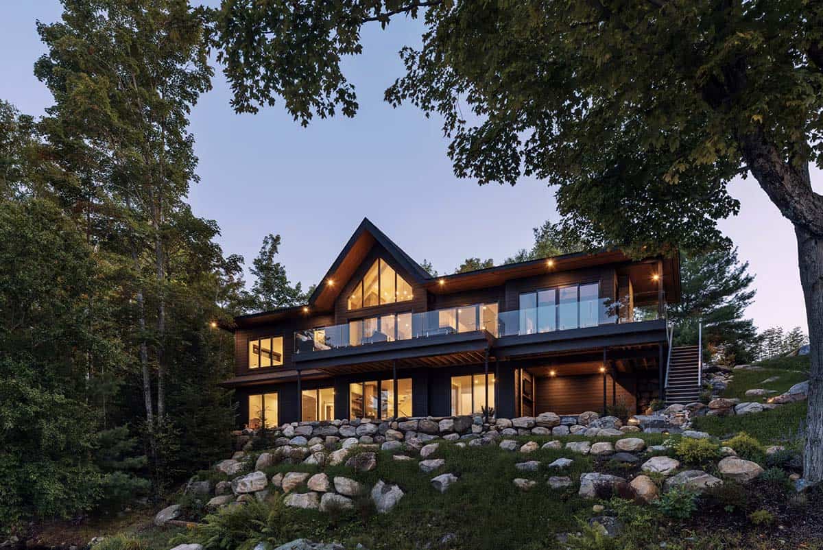 Two-story dark cottage at twilight with glowing gabled windows, glass balcony, and stone retaining wall