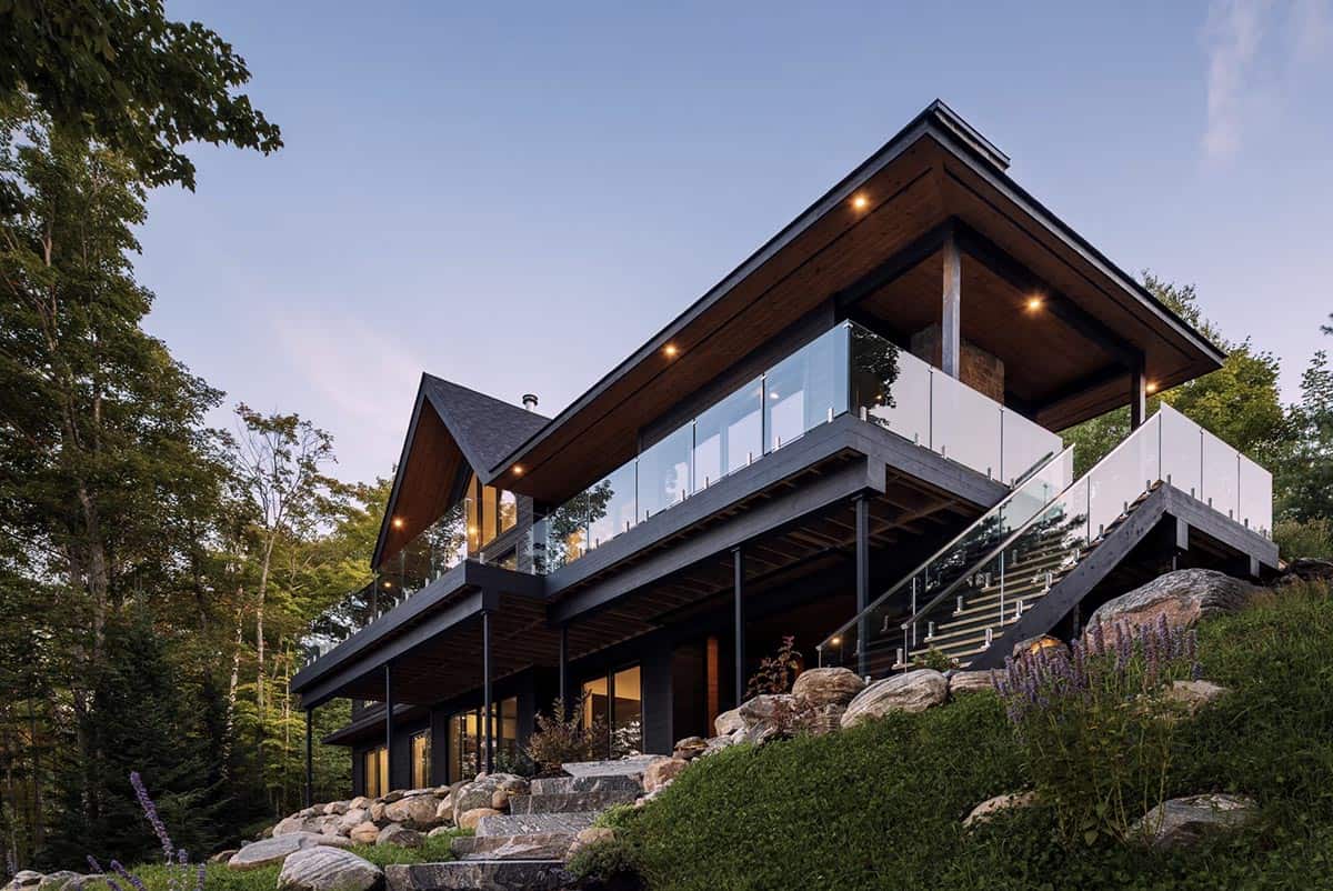 Cottage lakeside facade at dusk with cantilevered deck, glass railings, and wood soffit lighting