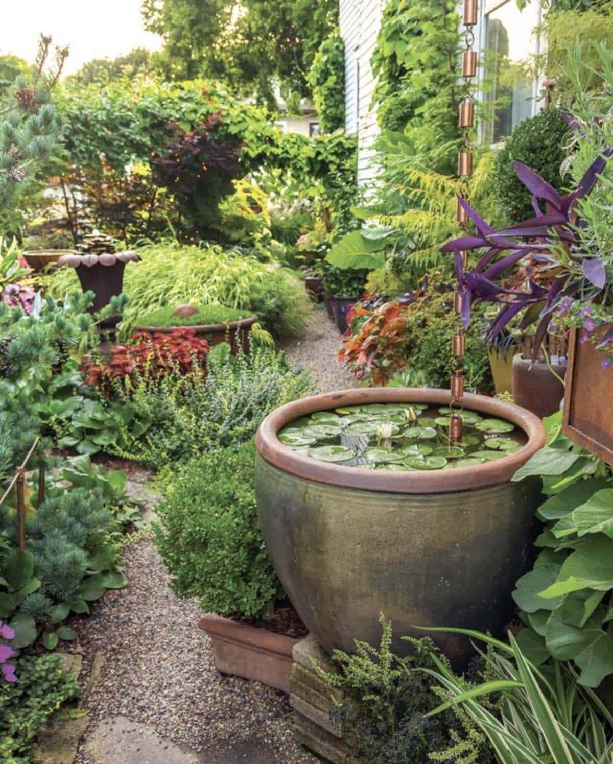 Narrow garden path with a large ceramic container pond, water lilies, copper rain chain, and lush container plantings