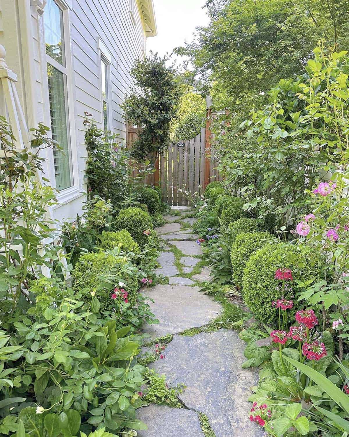 Narrow flagstone pathway lined with boxwood, pink flowers, and lush cottage garden plantings beside a gray house and wood gate