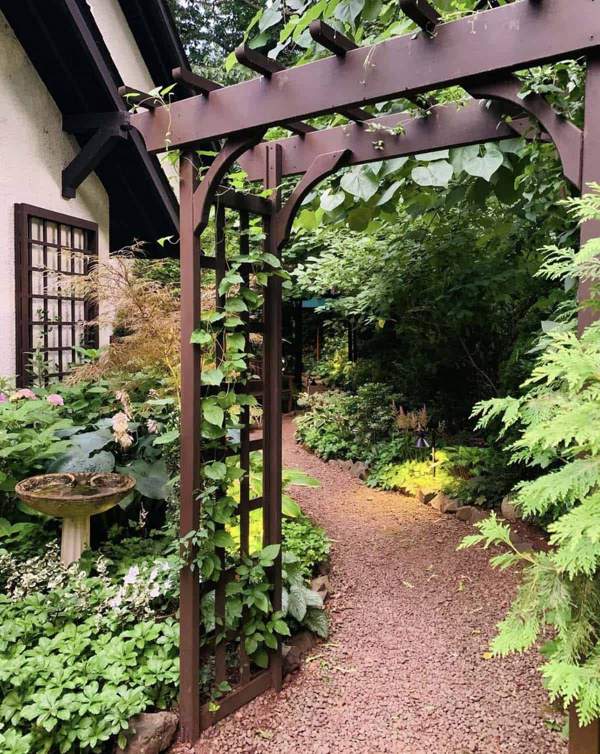 Narrow gravel path with a dark metal arbor, climbing vines, birdbath, hostas, ferns, and landscape lighting in a shaded garden