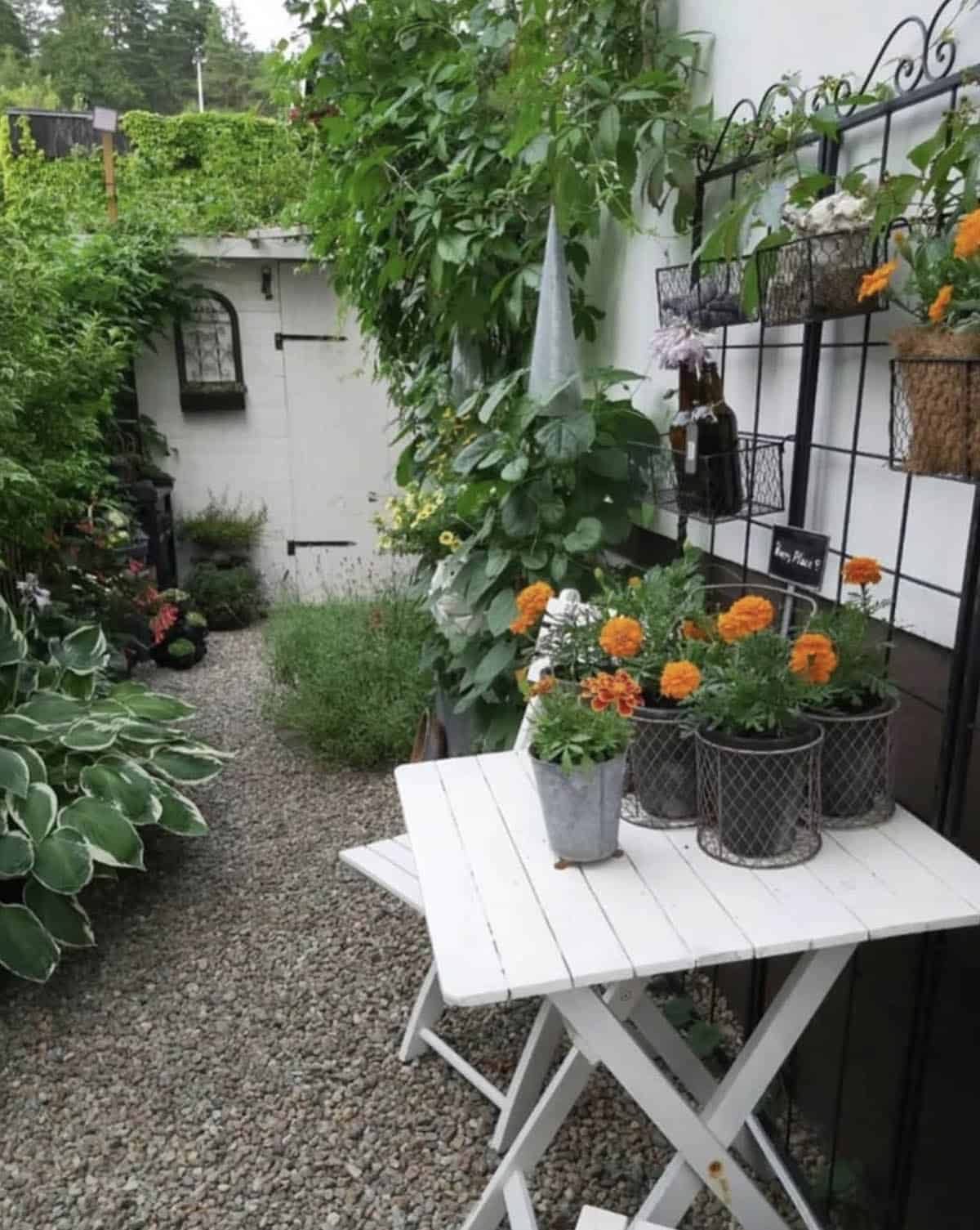 Narrow gravel side yard with a white bistro table, orange marigolds in galvanized pots, wall-mounted wire planters, hostas, and climbing vines