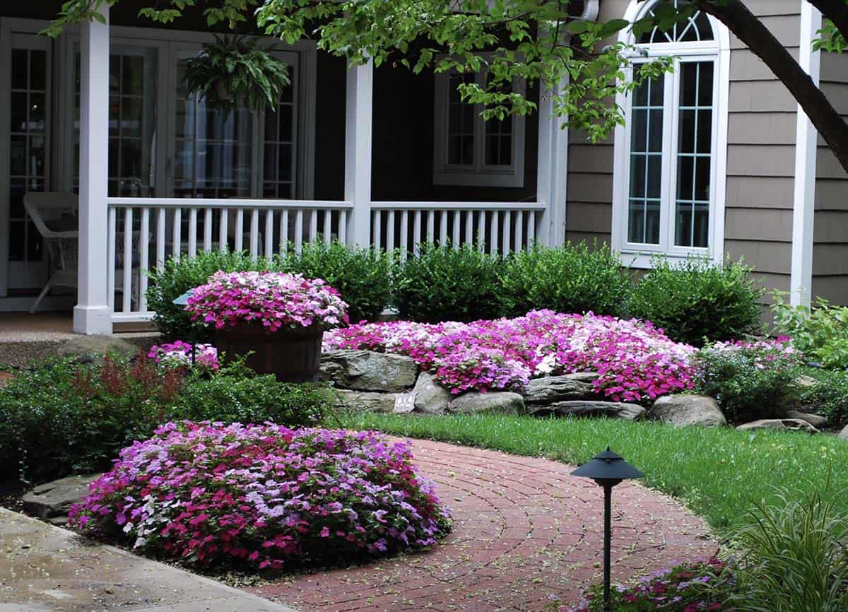 Small front yard landscaping with pink impatiens, stone borders, brick pathway, and white porch