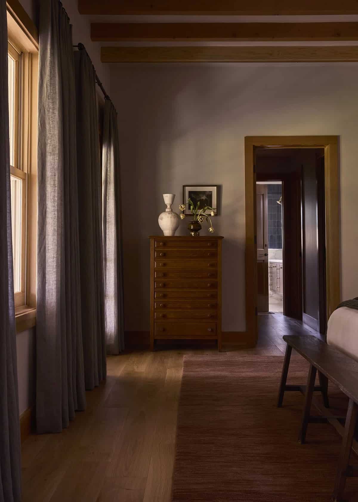 Primary bedroom with antique dresser, linen curtains, and view to en-suite bathroom