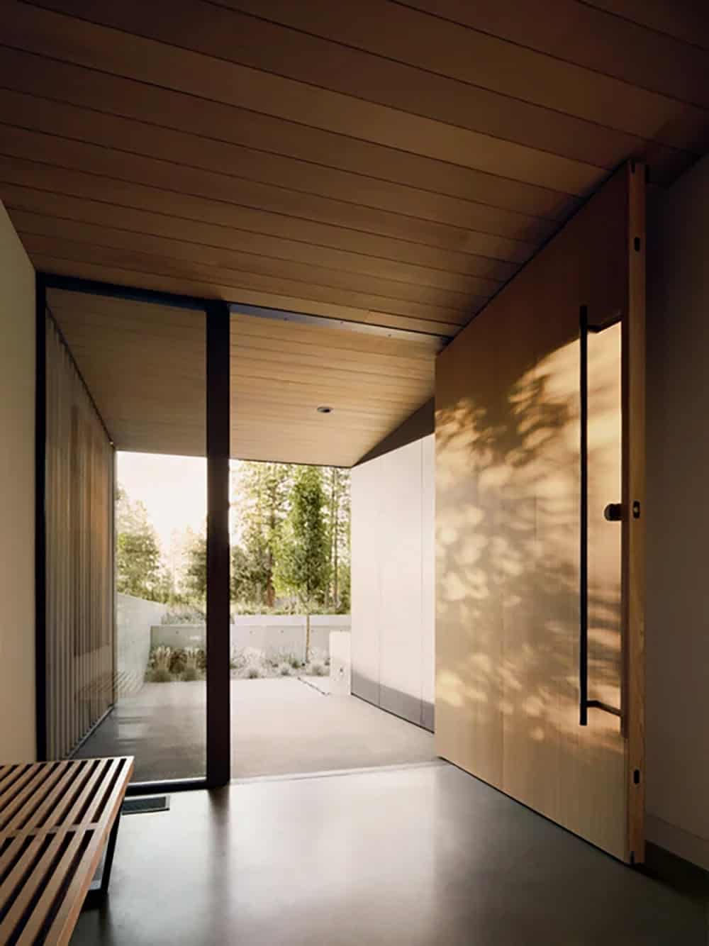 Minimalist wood-paneled entry door with black handle and slatted wood ceiling casting tree shadows