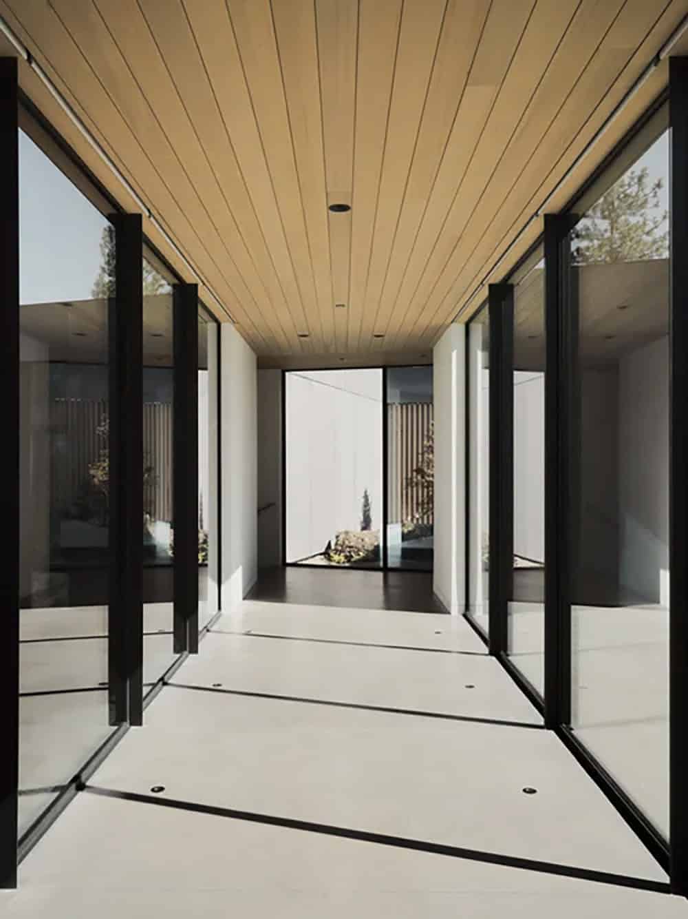 Symmetric glass-and-steel hallway with wood ceiling opening to a courtyard with ornamental plantings