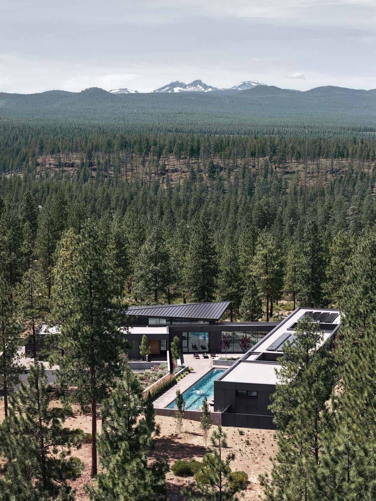 Aerial view of L-shaped modern home with lap pool set in vast Oregon pine forest with snowy Cascades beyond