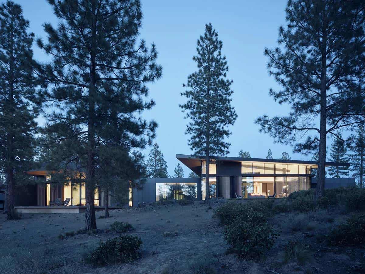 Contemporary home glowing warmly at twilight through floor-to-ceiling glass walls among pine trees