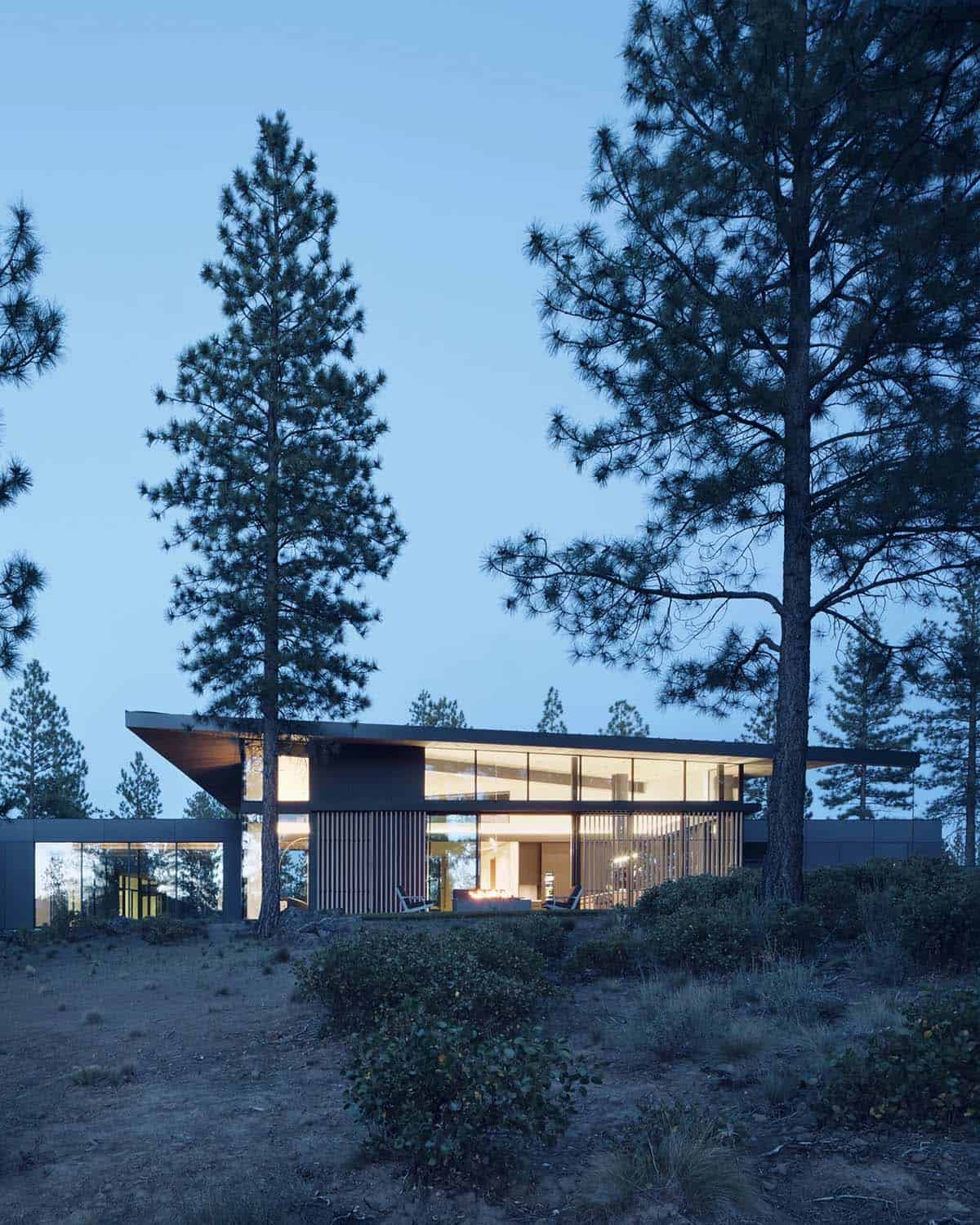 Illuminated modern home at twilight framed by tall ponderosa pines with cantilevered roofline and vertical wood siding