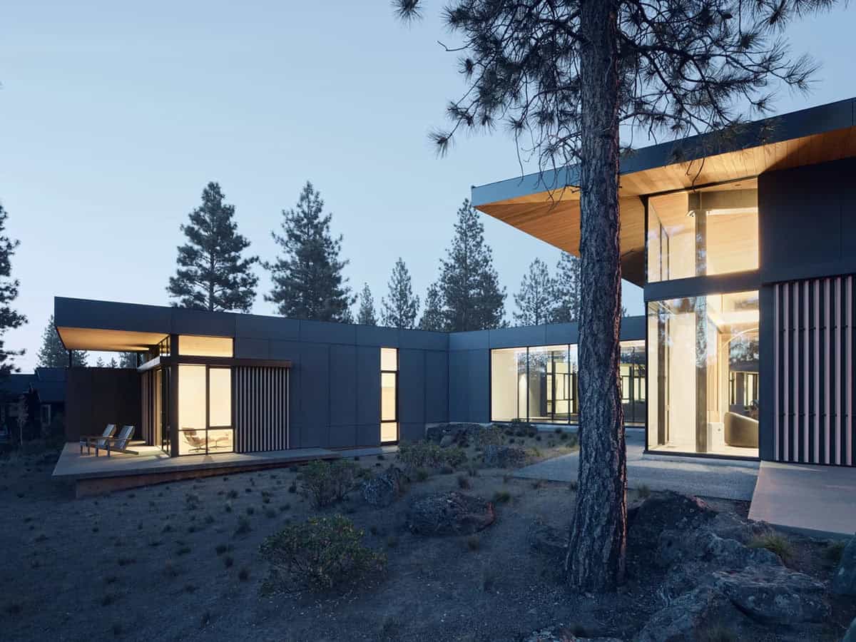 Angled exterior view of dark modern home at dusk showing two wings, wood-ceiling overhangs, and glowing interiors