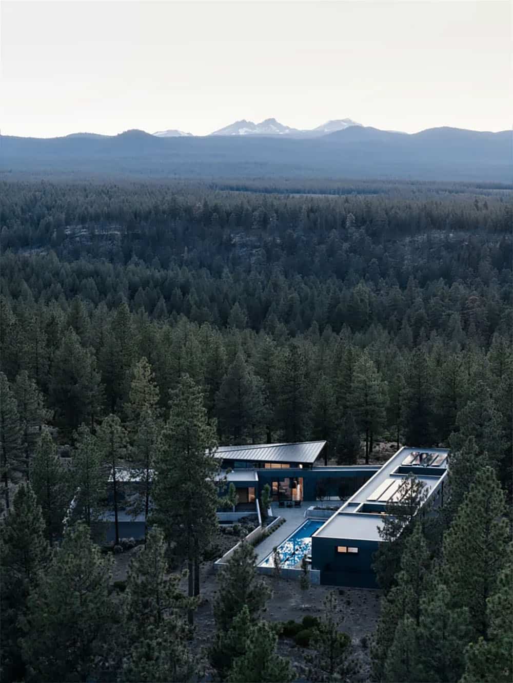 Aerial dusk view of L-shaped modern home with lap pool set within vast Oregon pine forest and Cascade Mountains beyond