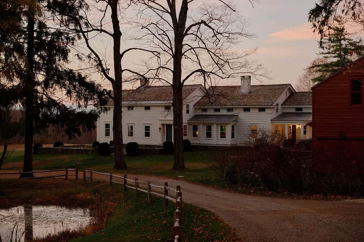 White farmhouse exterior at dusk with gravel driveway, pond, split-rail fence, and red barn