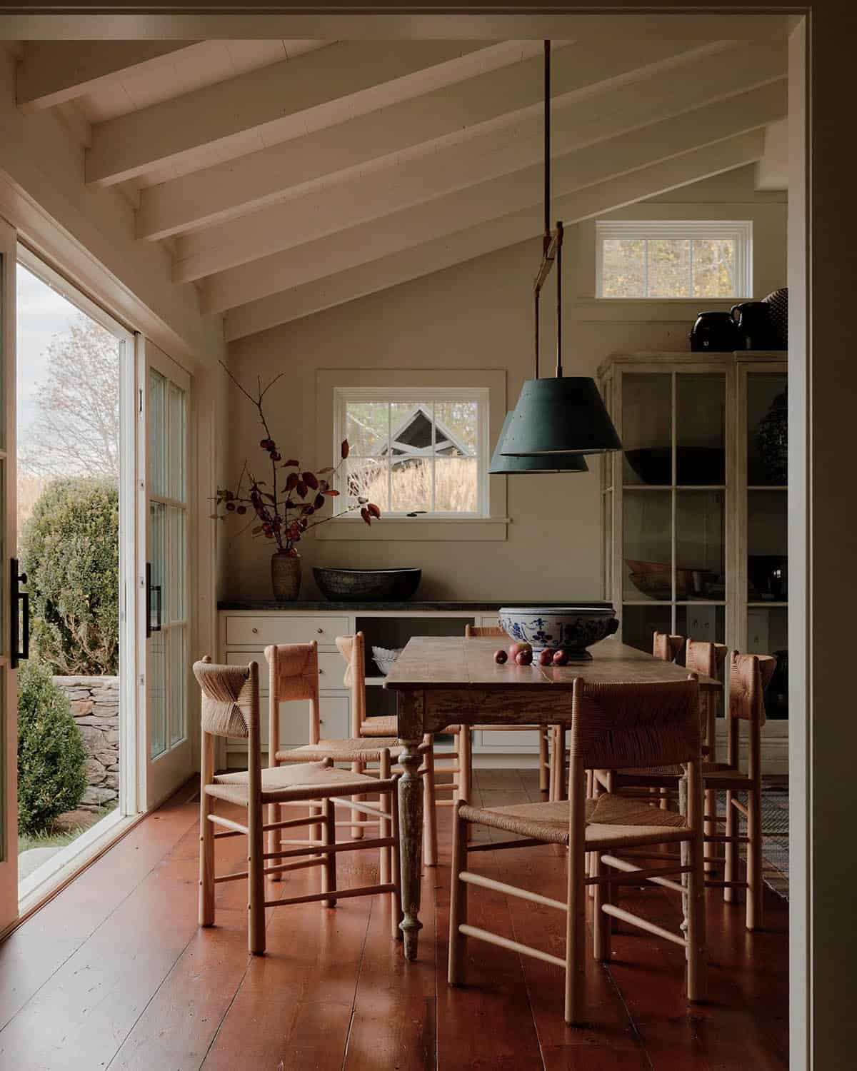 Dining area with rush-seat chairs around an antique table, cathedral ceiling with exposed beams, and garden doors