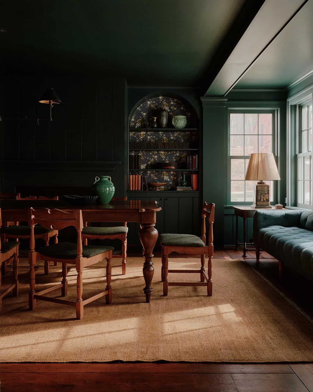 Dark green dining room with antique wood table, upholstered chairs, arched bookcase, and tufted settee