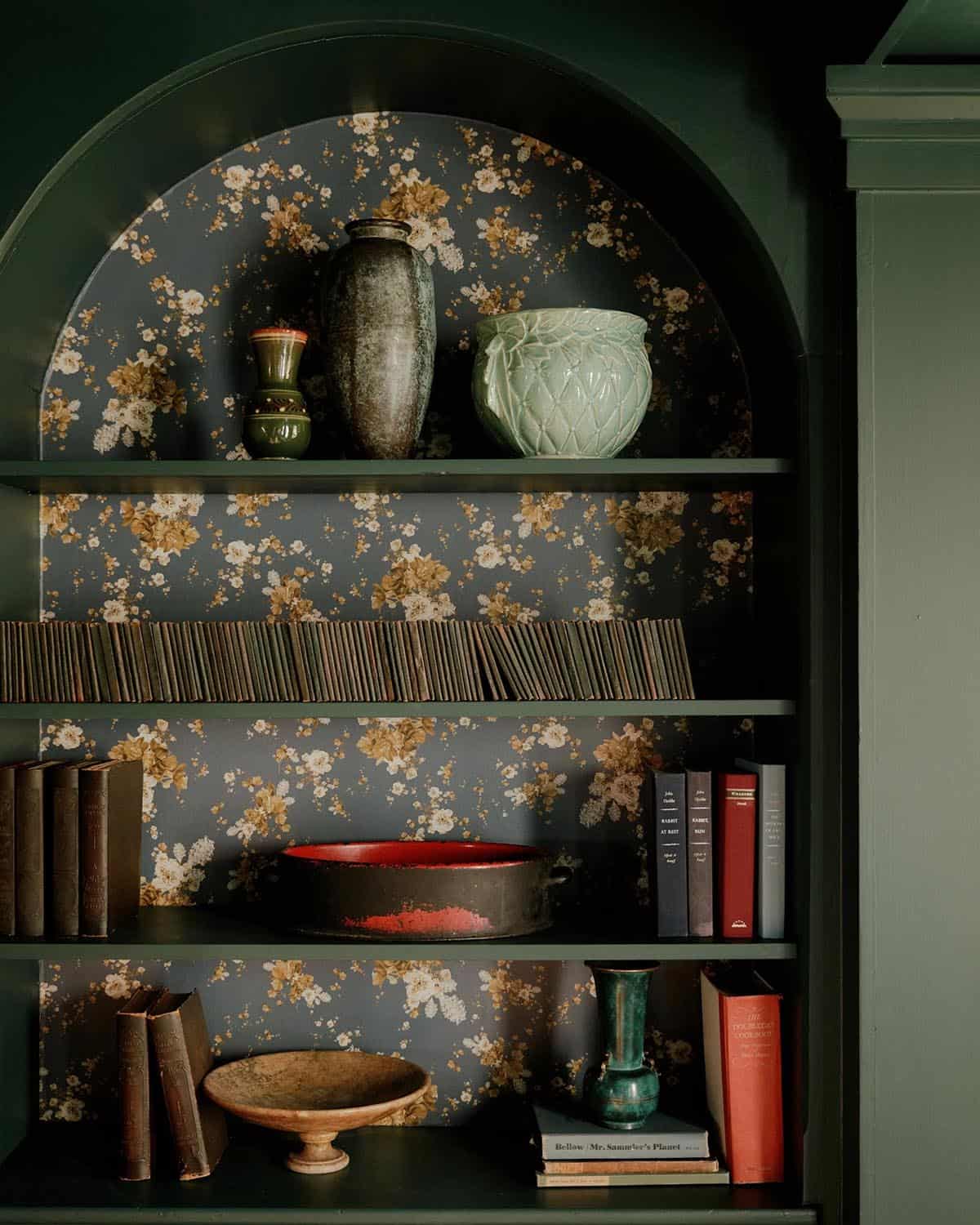 Arched built-in bookshelf lined with dark floral wallpaper, ceramic vessels, and vintage books