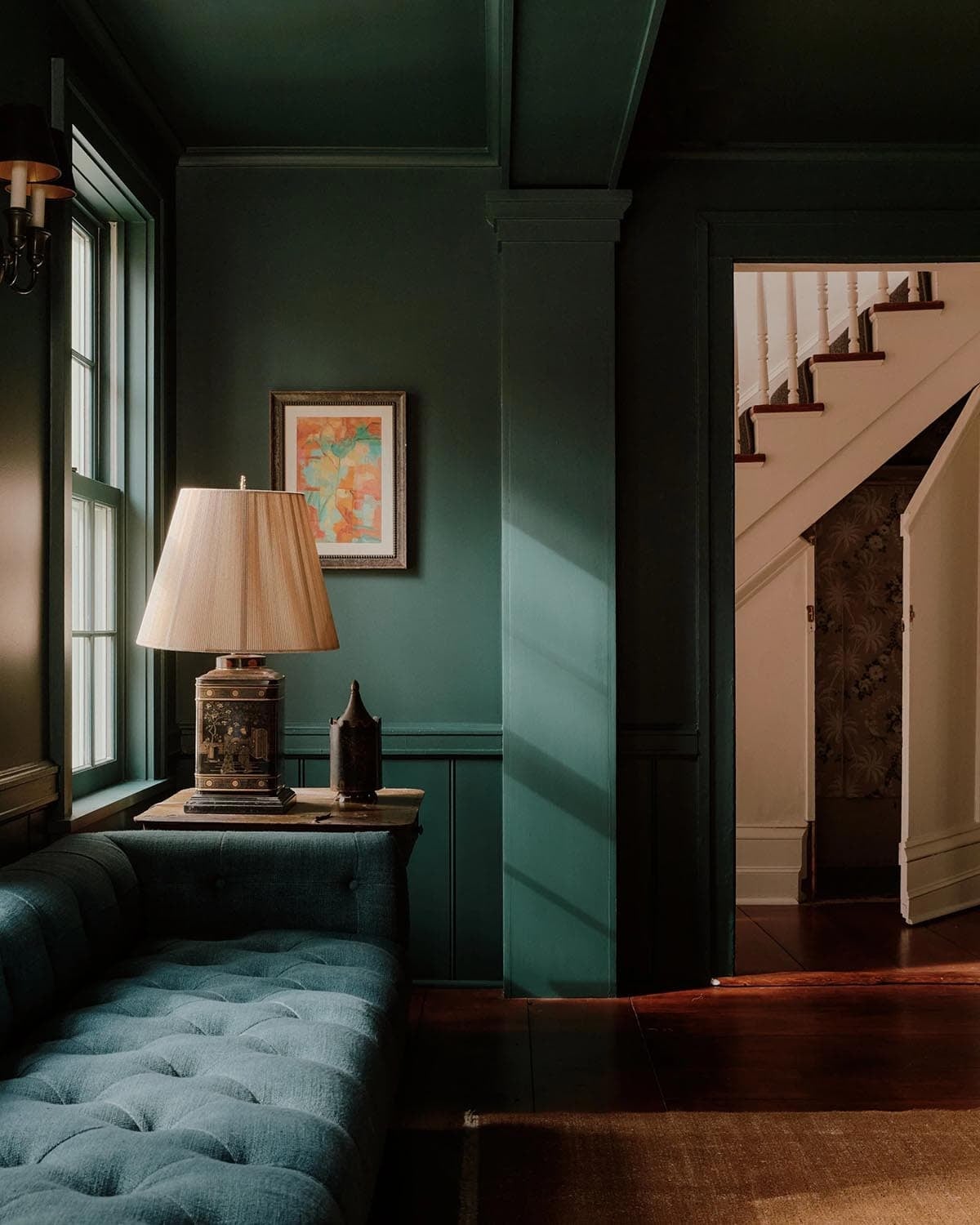 Dark green parlor corner with tufted sofa, table lamp, colorful abstract painting, and staircase beyond