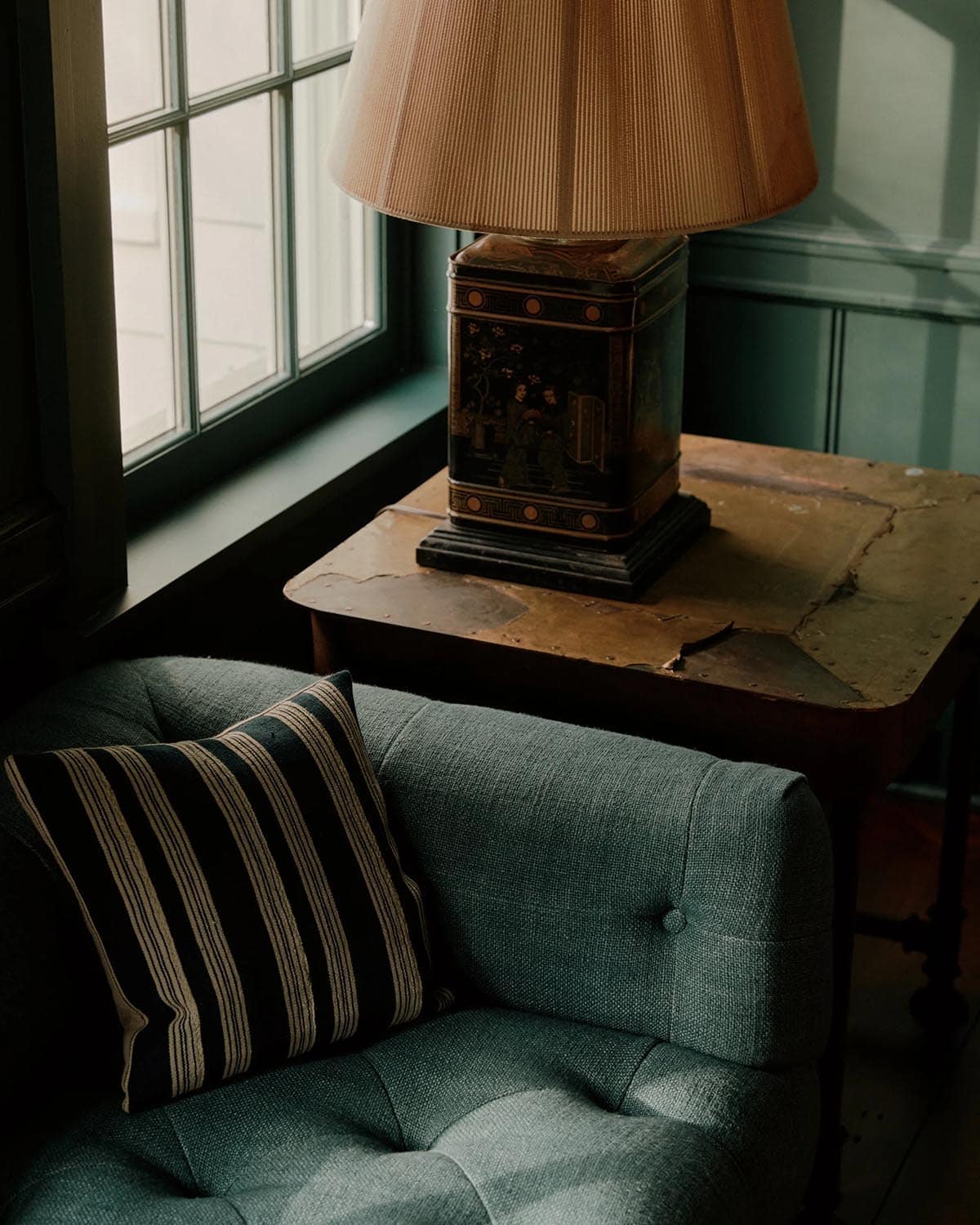 Close-up of tufted blue-green sofa arm with striped pillow and antique chinoiserie tea caddy lamp on worn side table