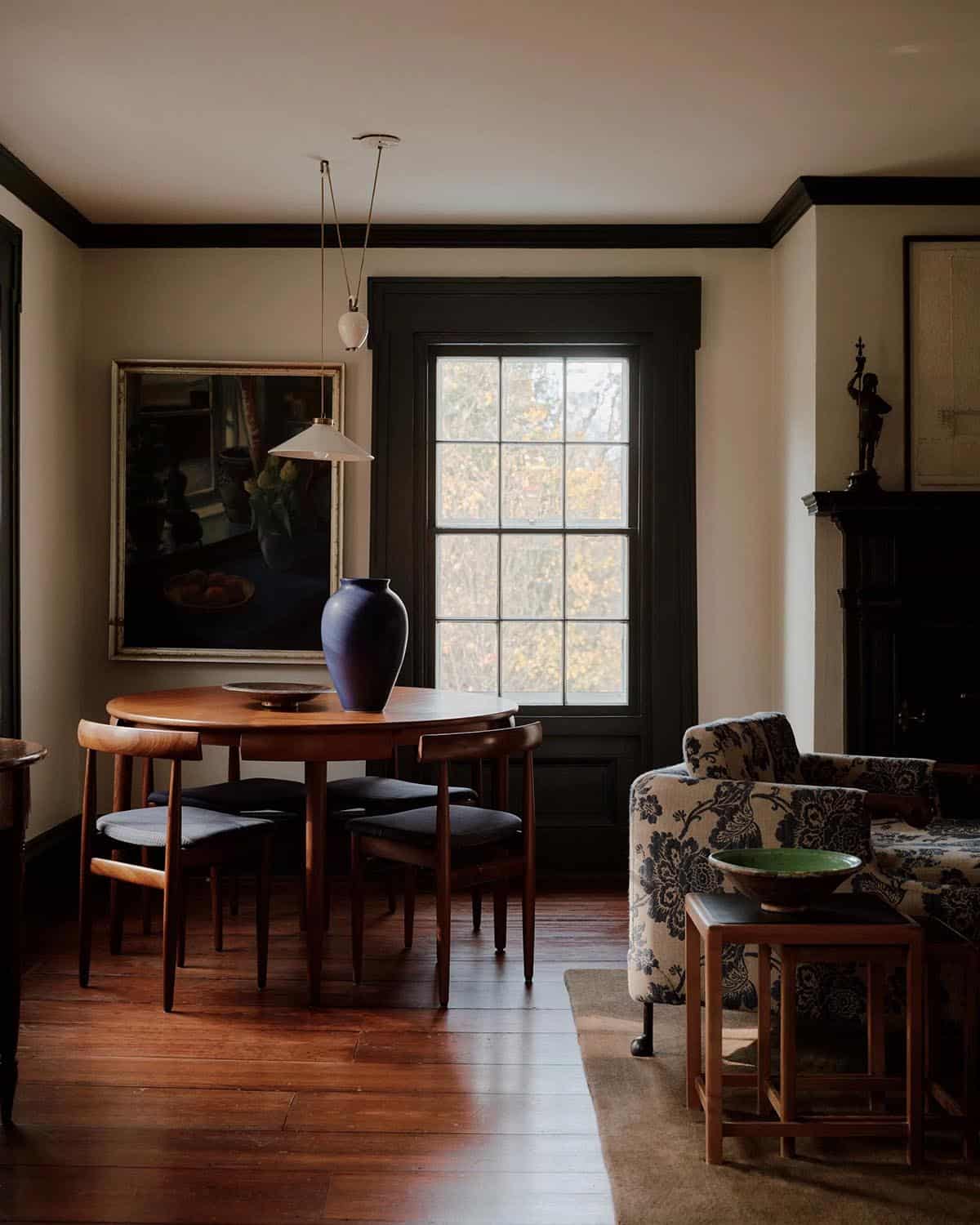 Living-dining room with mid-century round table, walnut chairs, blue-floral armchair, and dark window trim