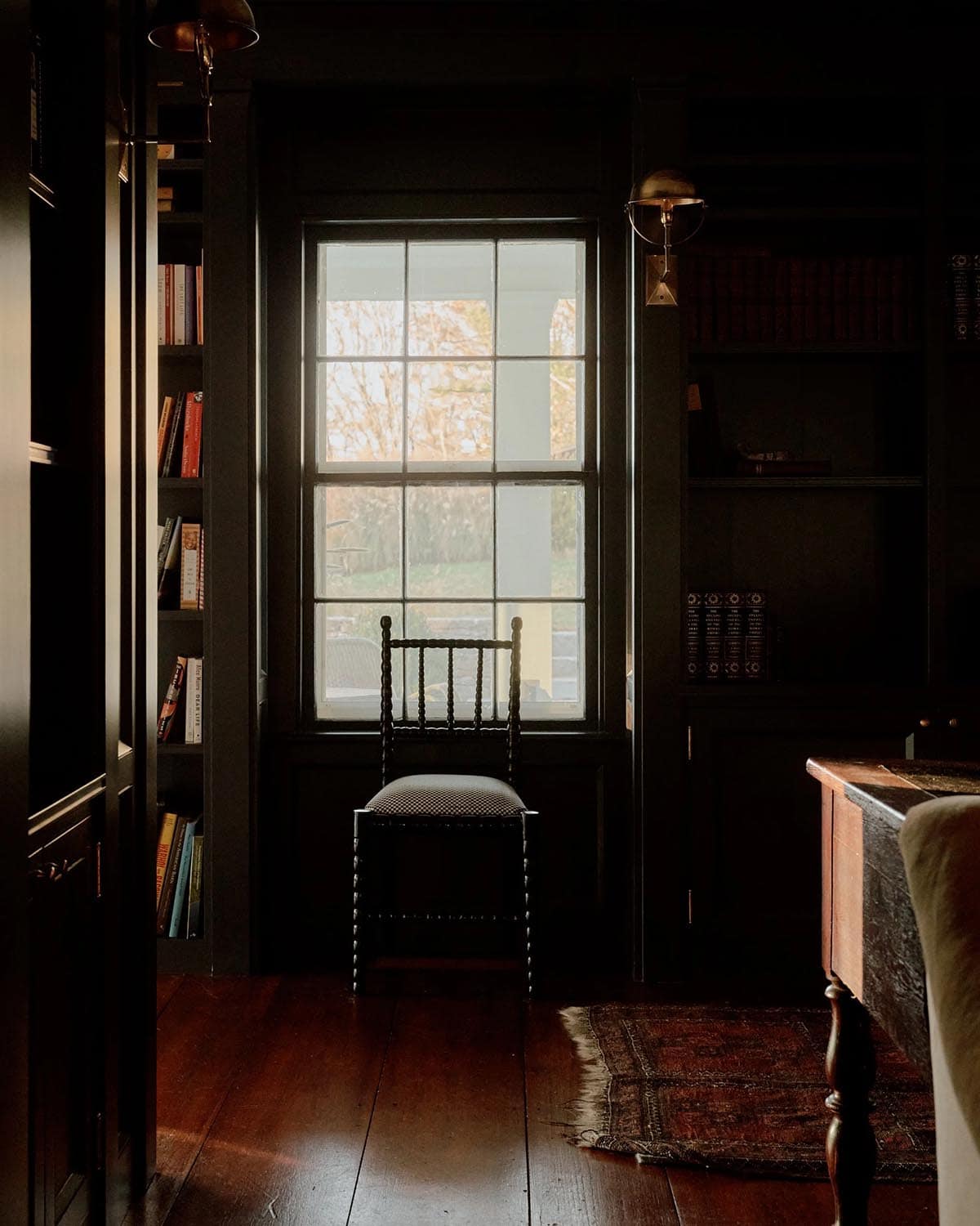 Dark moody study with floor-to-ceiling bookshelves, single chair silhouetted against tall window