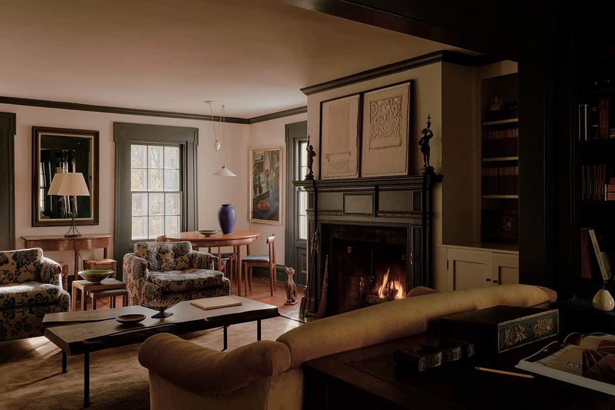 Living room with lit fireplace, ornate mantel, floral armchairs, mid-century dining table, and built-in shelving