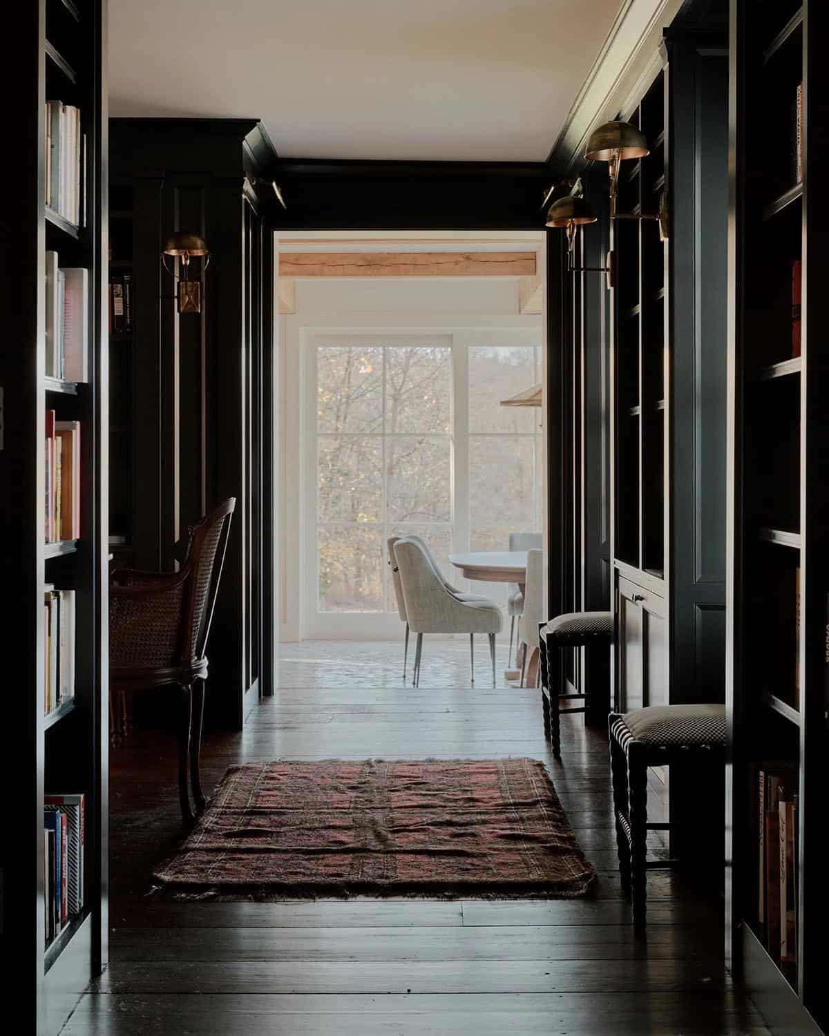 Dark library hallway lined floor-to-ceiling with books and brass dome sconces, opening to bright room beyond