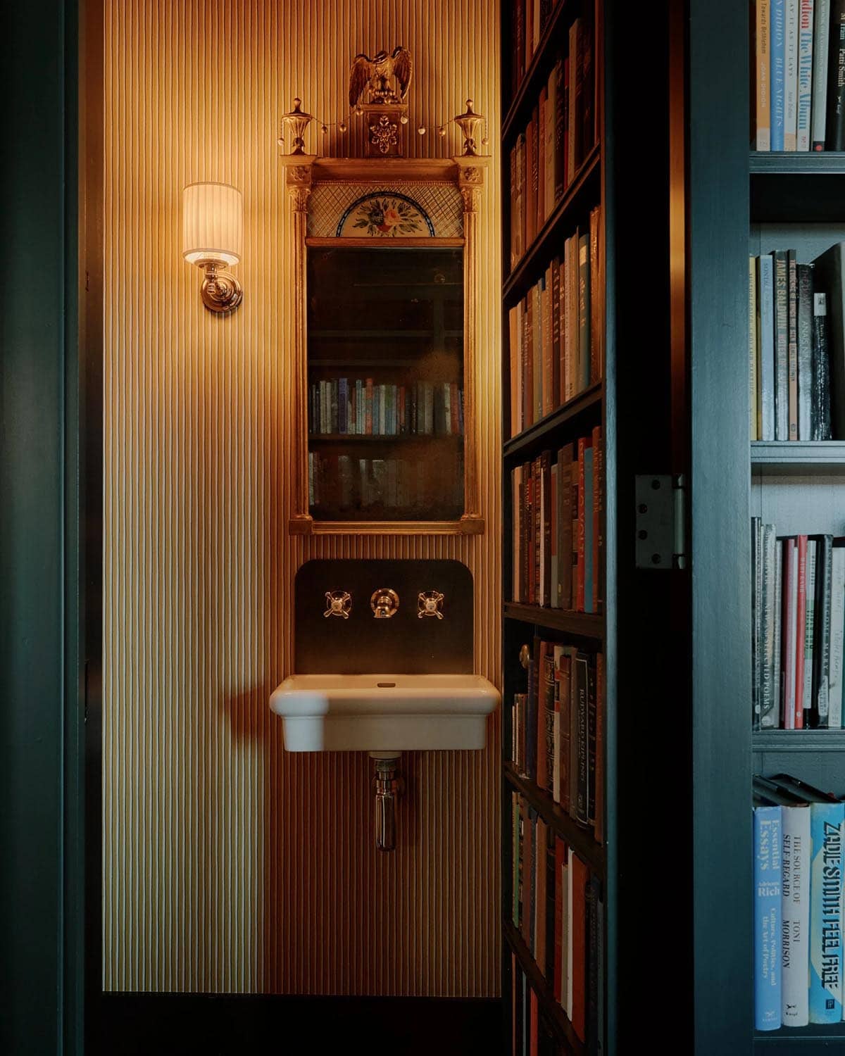 Hidden powder room revealed through bookcase door, with gold striped wallpaper, pedestal sink, and ornate antique mirror