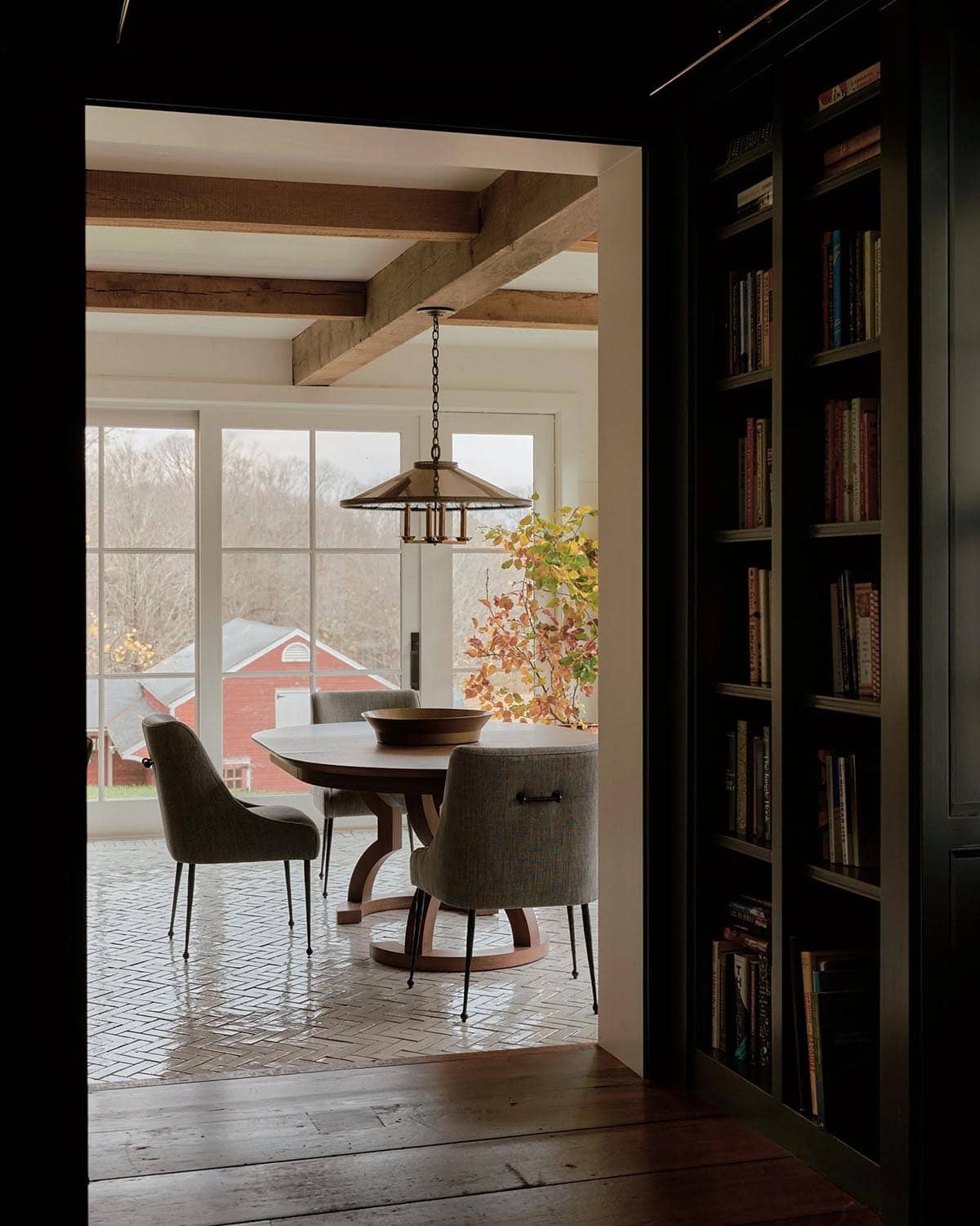 View from dark library into bright breakfast nook with round pedestal table, upholstered chairs, herringbone tile floor, and exposed wood beams