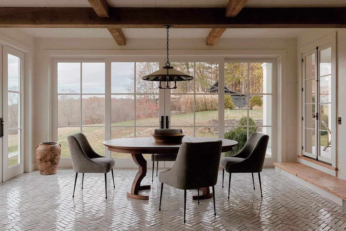 Sunlit breakfast nook with round walnut table, gray upholstered chairs, herringbone tile floor, exposed beams, and panoramic garden views