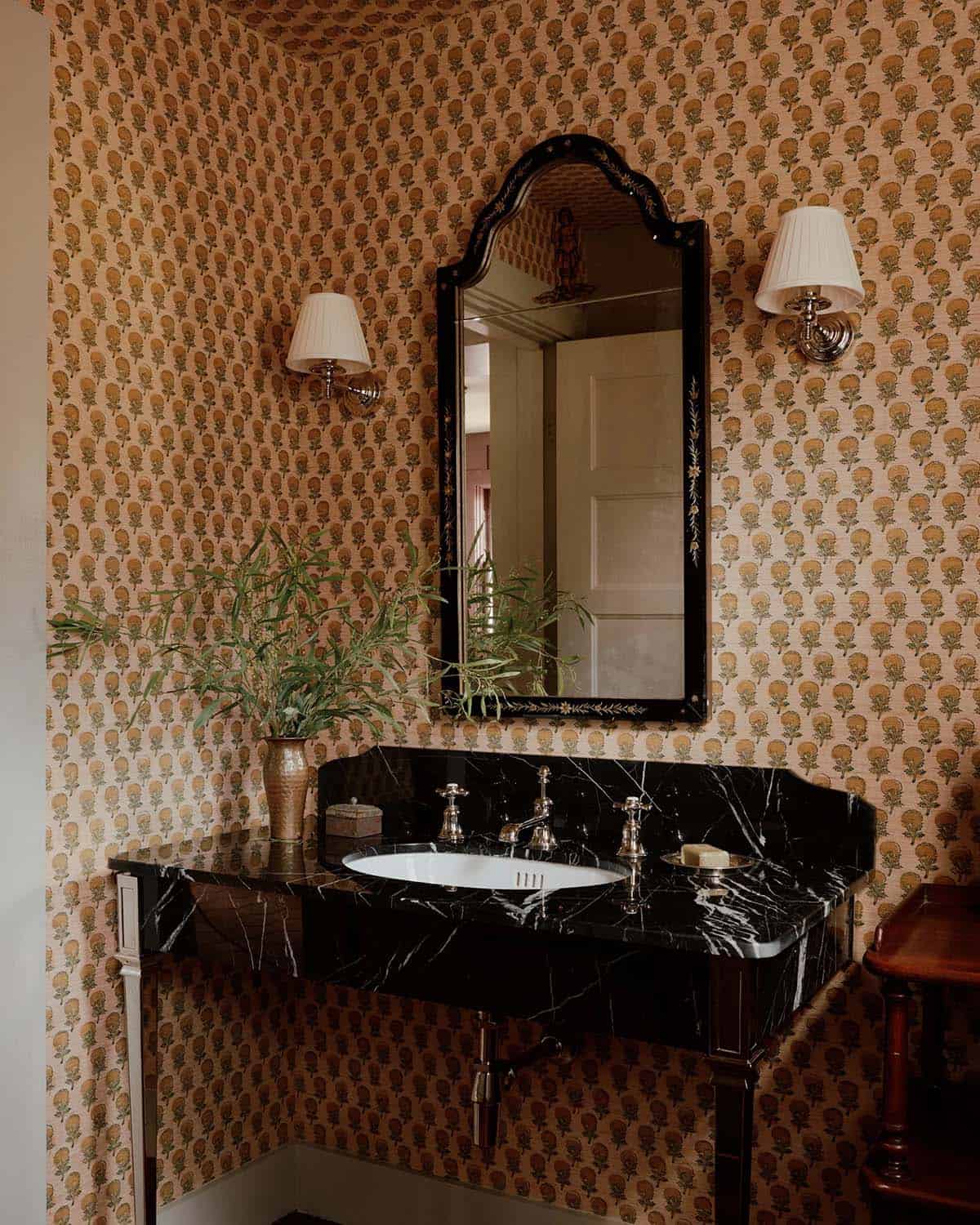 Powder room with warm floral block-print wallpaper, black marble vanity, ornate arched mirror, and chrome sconces