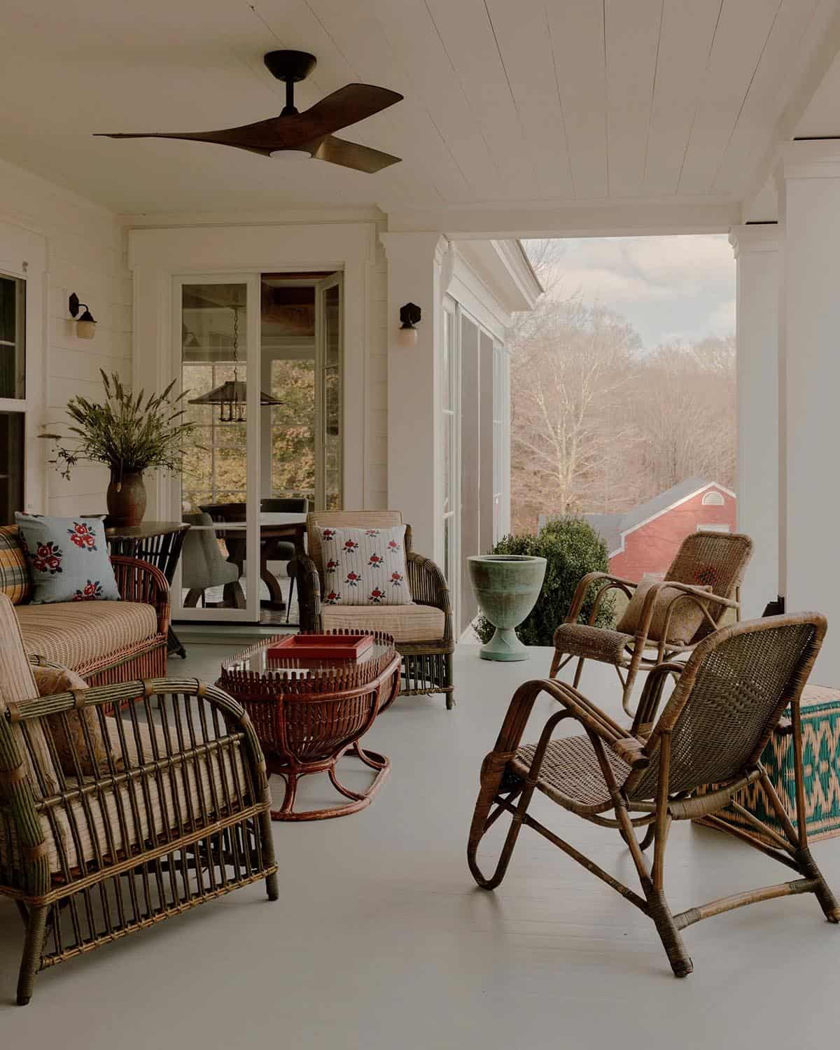 Covered porch with mixed vintage rattan and wicker seating, floral pillows, ceiling fan, and view of red barn