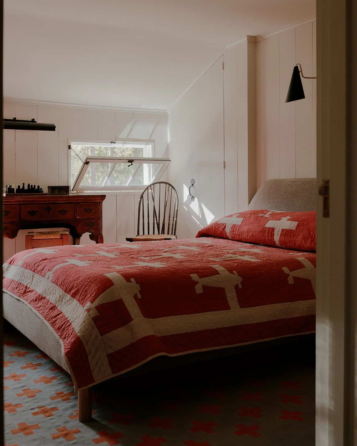 Bright bedroom with red and white cross-pattern quilt, antique mahogany dresser, Windsor chair, and cross-motif rug