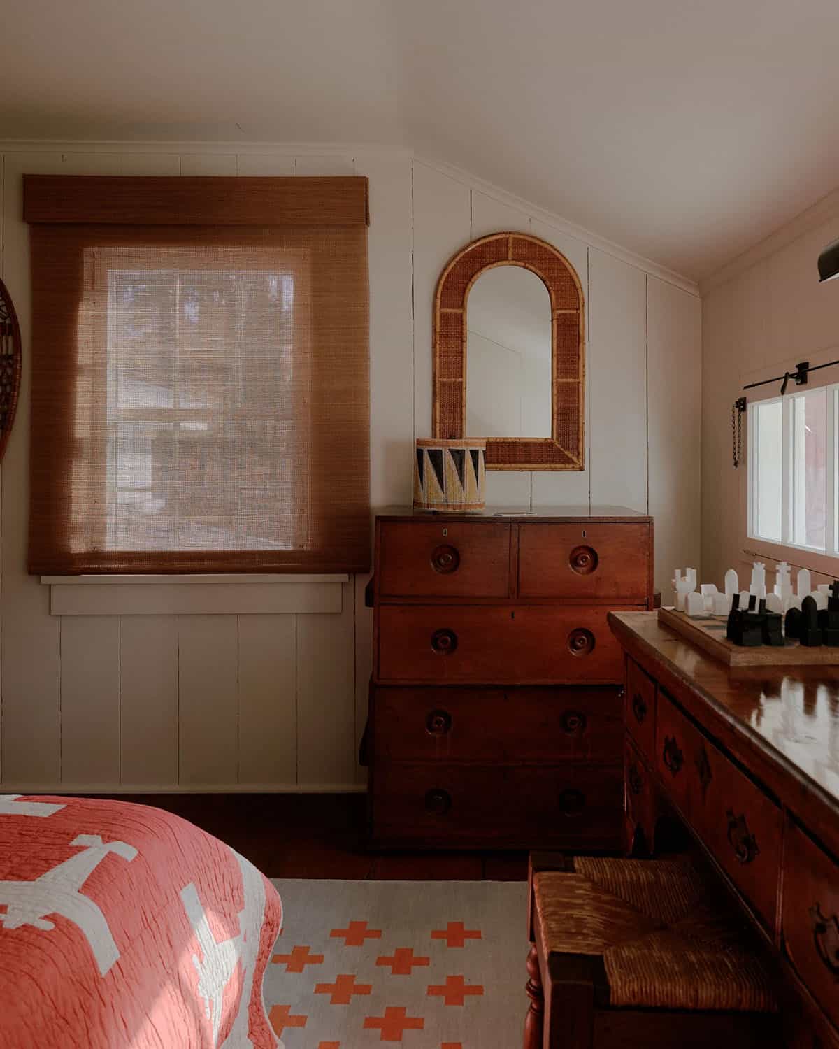 Bedroom corner with antique red dresser, arched rattan mirror, wicker Roman shade, and chess set on side table
