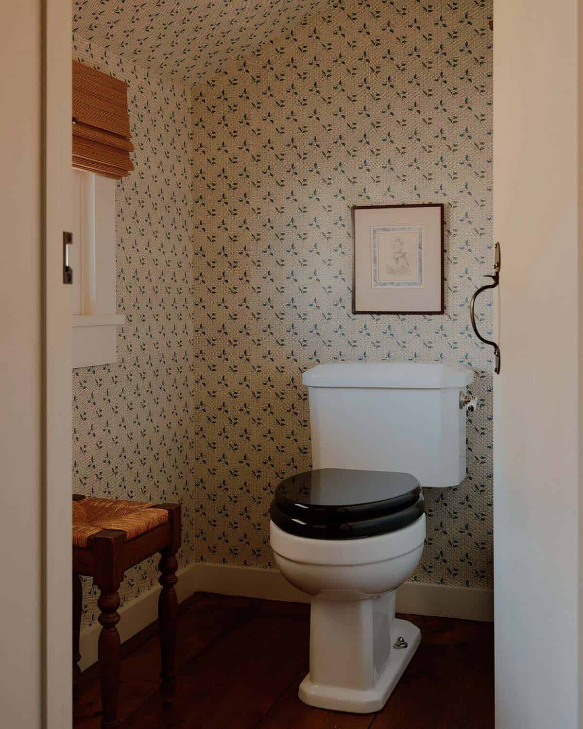 Compact bathroom with delicate sprig wallpaper, black lacquer toilet seat, rush stool, and framed artwork