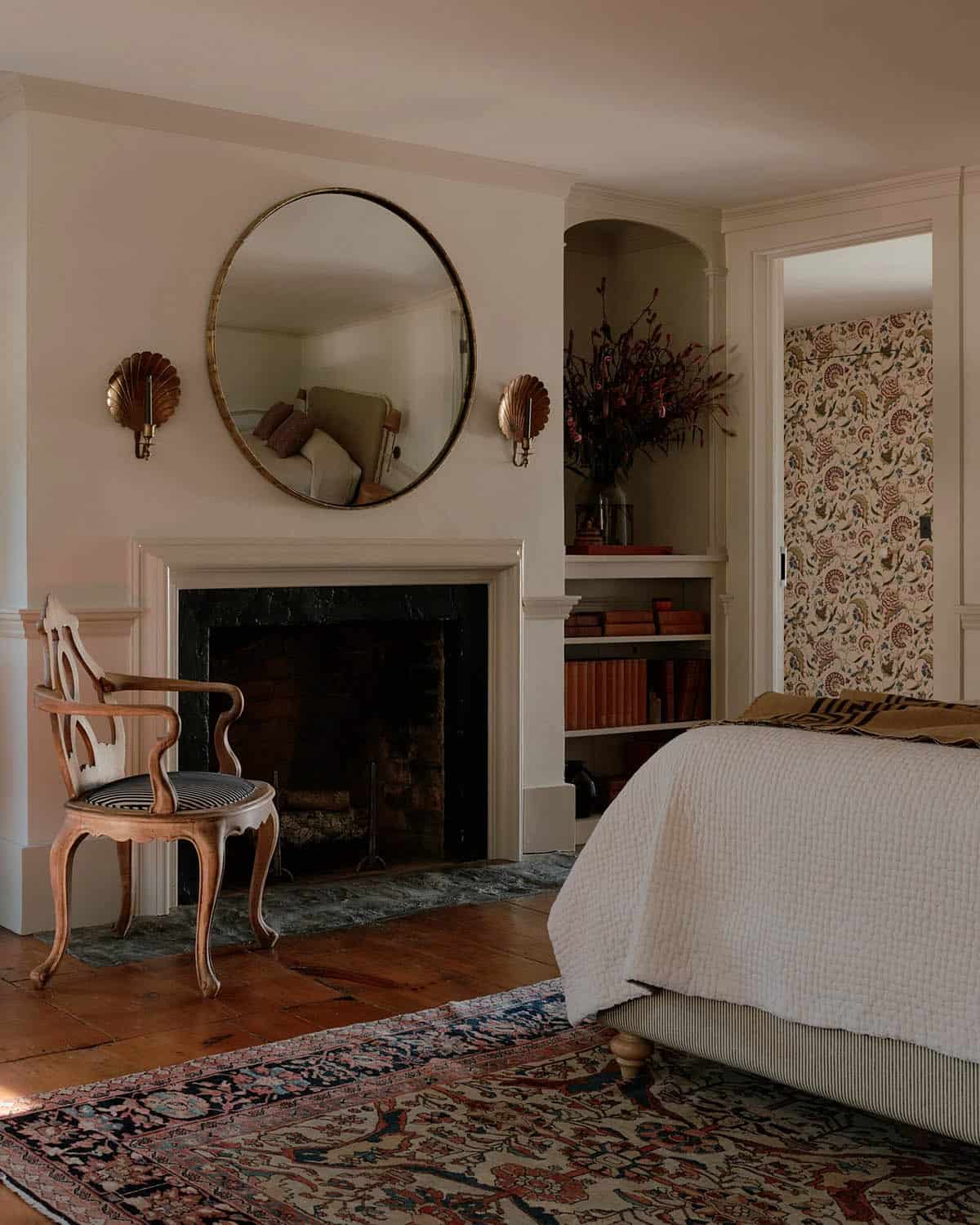 Primary bedroom fireplace wall with brass round mirror, shell sconces, ornate carved chair, Persian rug, and botanical wallpaper beyond doorway