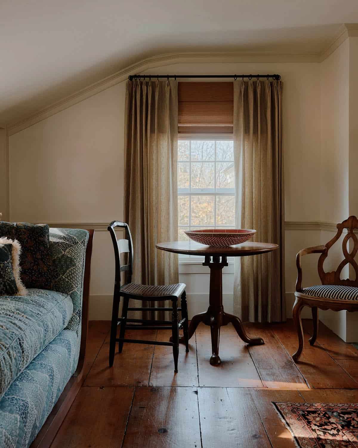 Bedroom sitting area with pedestal table, rush-seat chair, carved armchair, striped drapes, and wide-plank floors