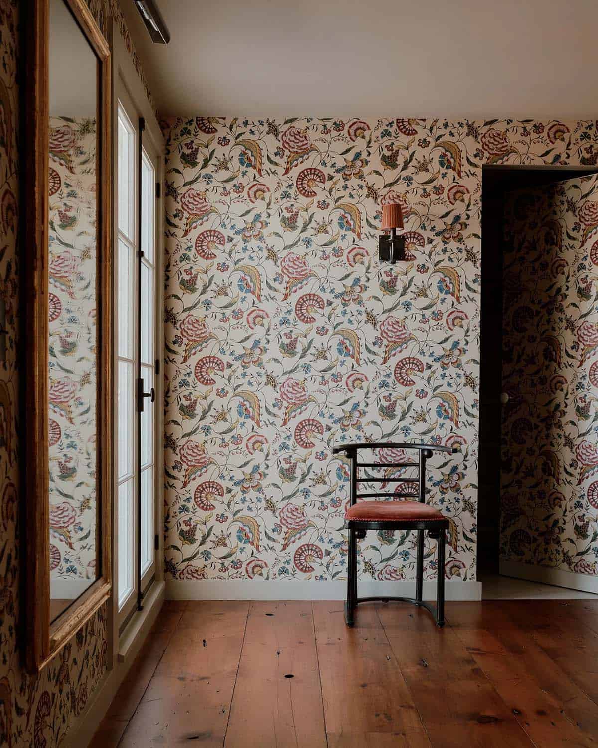 Hallway corner covered floor-to-ceiling in bold tropical crewelwork wallpaper with a single black chair and sconce