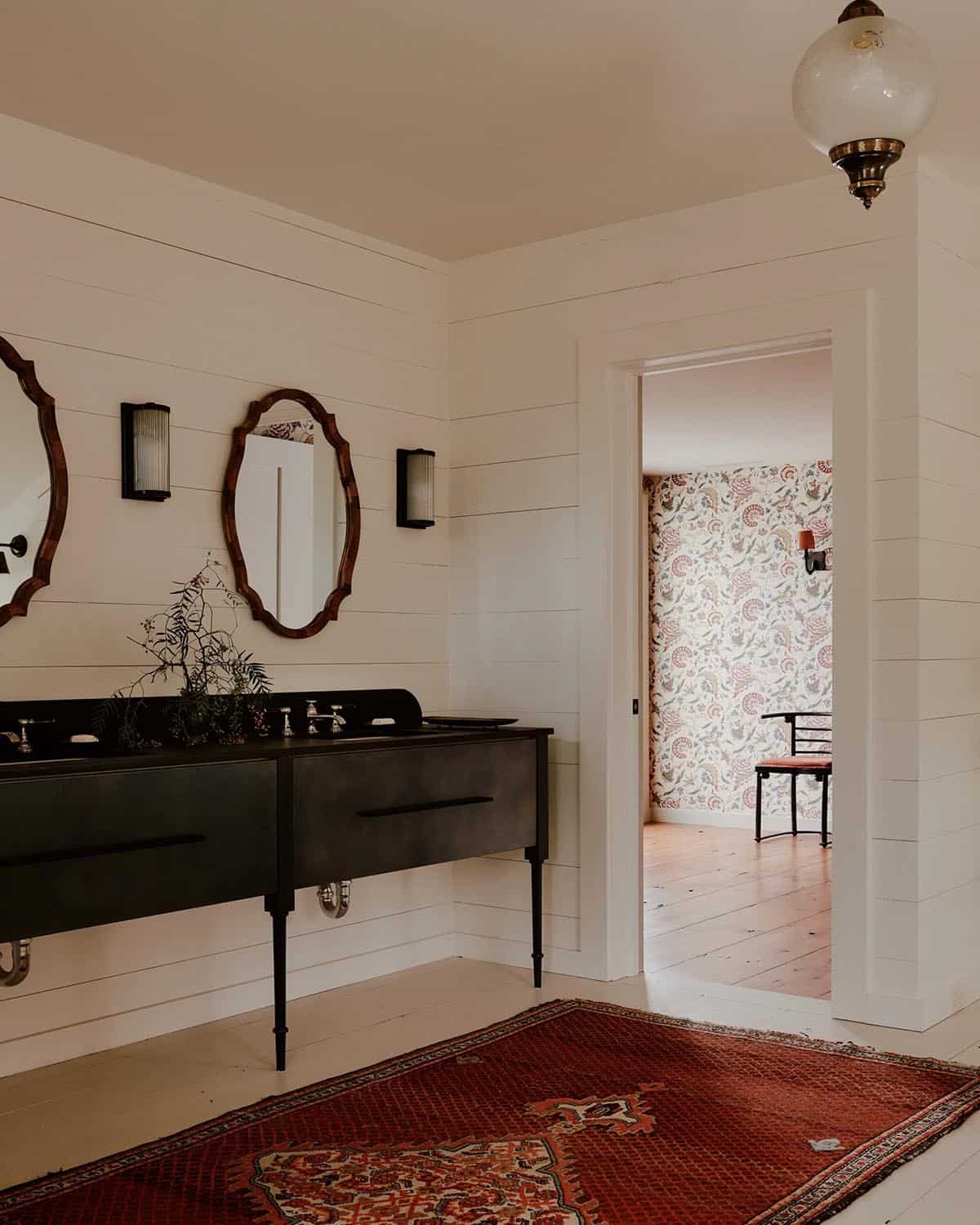 Bathroom with dark steel double vanity, carved wood oval mirrors, globe pendant, red Persian rug, and view into wallpapered room