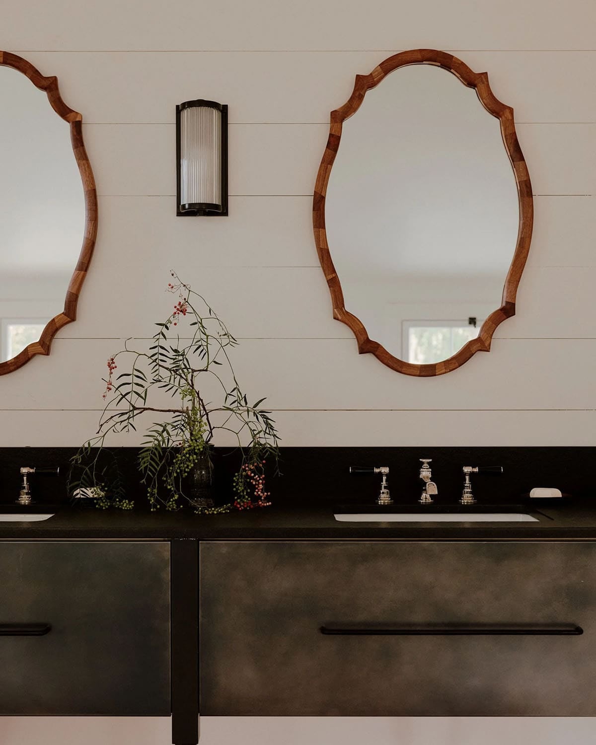 Close-up of double bathroom vanity with dark steel cabinet, black stone countertop, chrome fixtures, wood-framed oval mirrors, and botanical stem arrangement