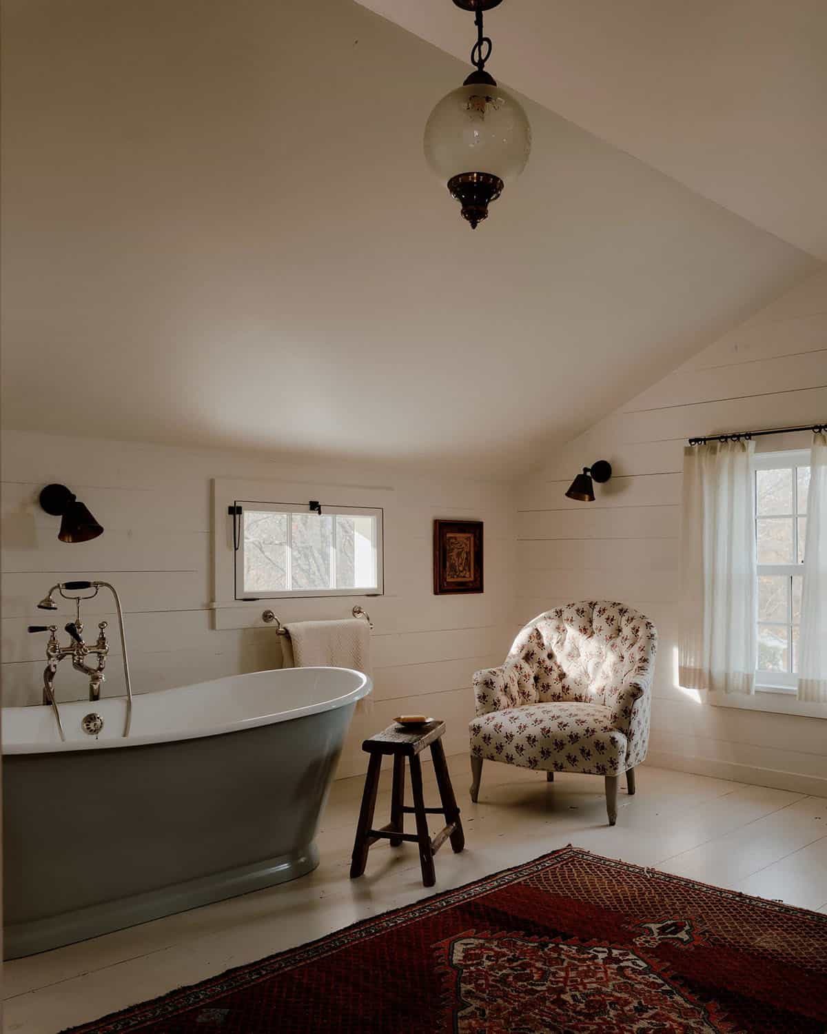 Spacious bathroom with freestanding gray slipper tub, floral armchair, Persian rug, globe pendant, and shiplap walls