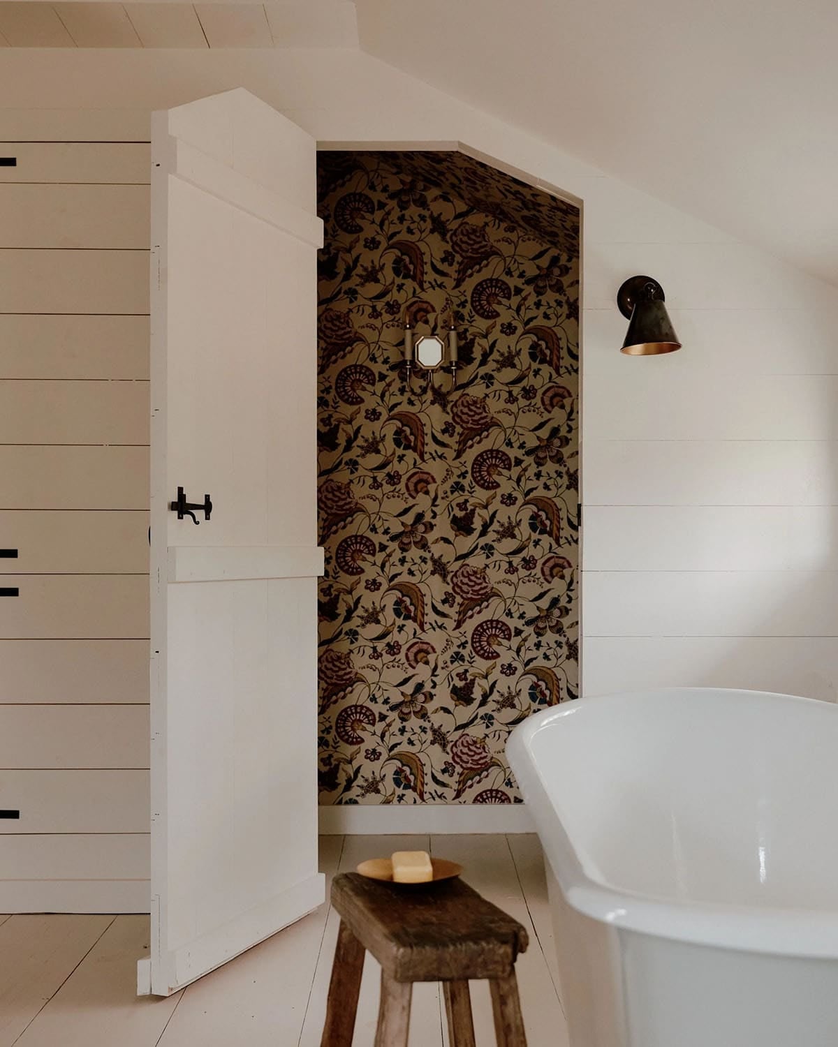 Bathroom nook with bold tropical crewelwork wallpaper visible through open Dutch door beside freestanding soaking tub